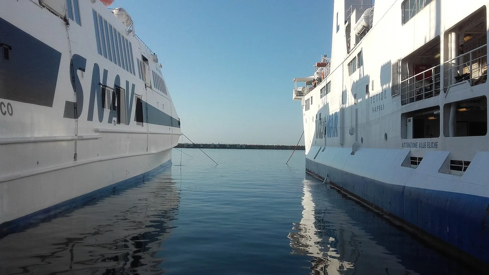 Due navi passeggeri ormeggiate una accanto all'altra nel porto di Formia, con l'acqua calma in mezzo e la diga foranea sullo sfondo